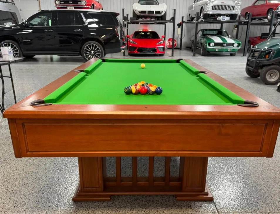 Oak pedestal pool table with green felt, racked and ready, in a showroom garage